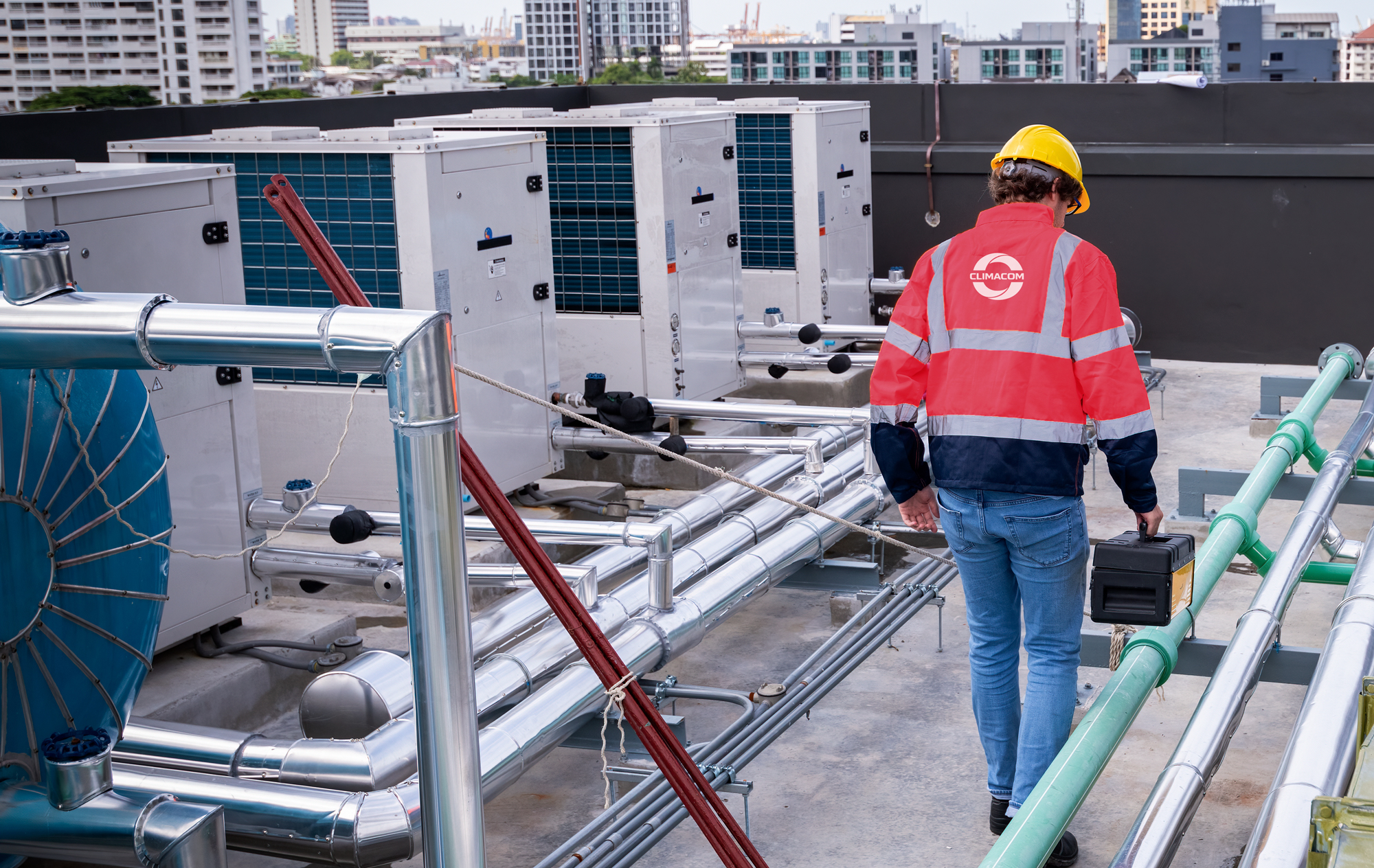 Commercial HVAC technician carrying tools through a rooftop plant area with multiple air conditioning units and service piping